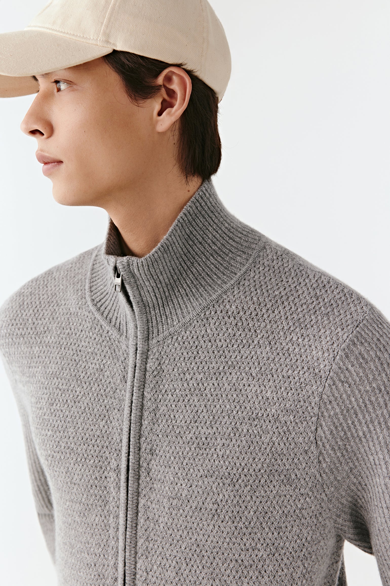 Person wearing a gray zip-up sweater and beige cap on a light background