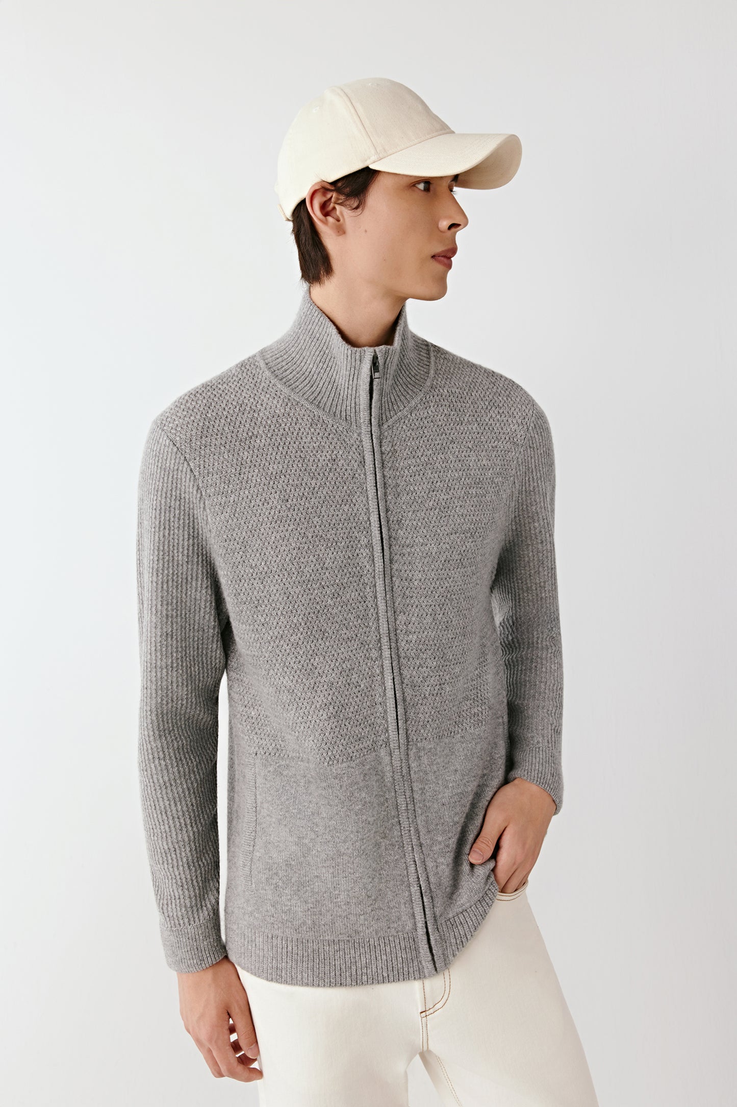 Person wearing a gray zip-up sweater and white cap on a light background