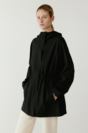 Full-length studio photograph of a woman wearing a black short trench coat in wool and silk with a relaxed oversized cut. The coat has a high collar, wide sleeves and elasticated details that gather the fabric slightly at the waist. It ends at hip length and is worn over light trousers.