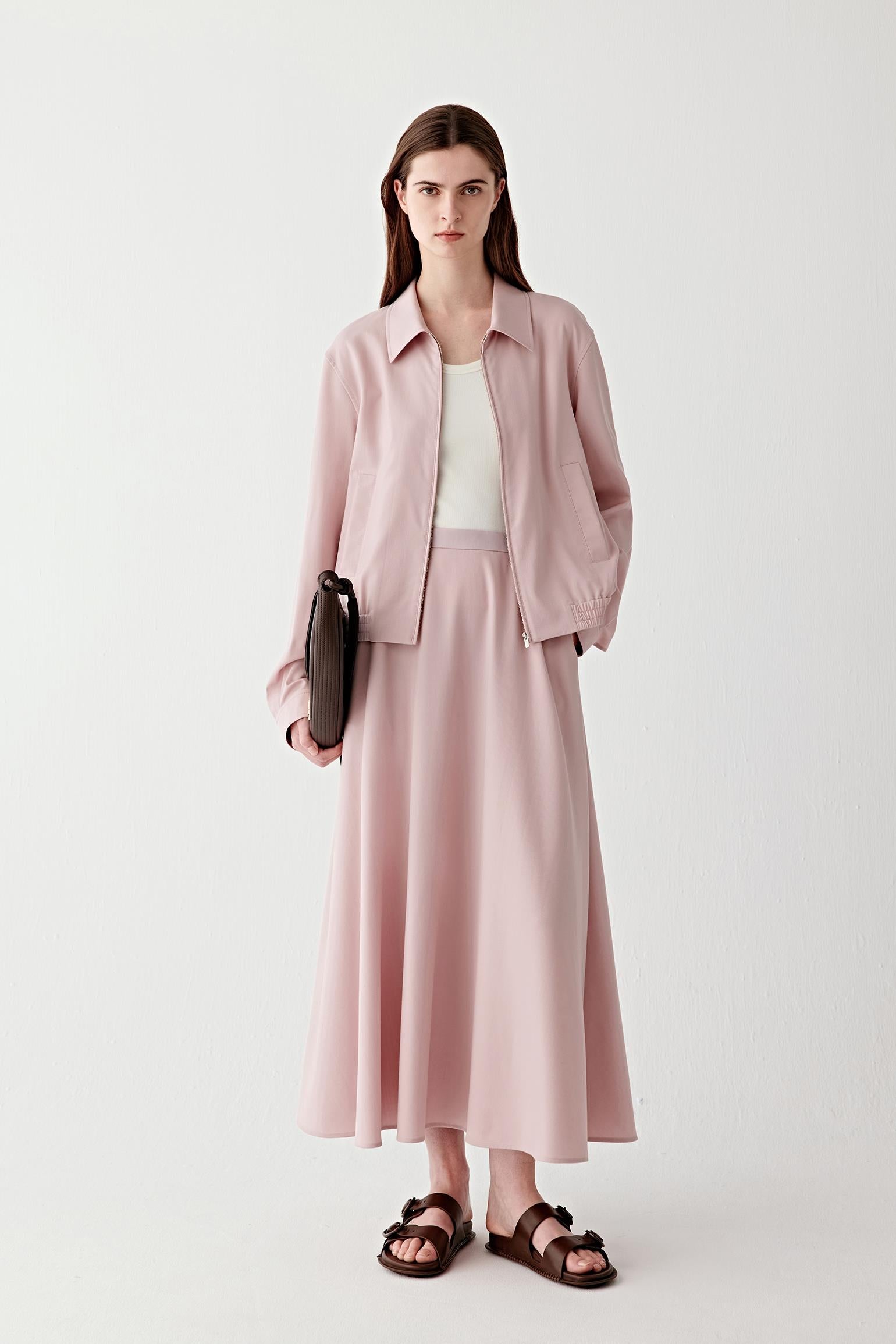 Full-length front view of a soft pink wool twill jacket worn open, featuring a straight cut, collarless neckline, long sleeves and a relaxed fit, styled with a light top and matching skirt.