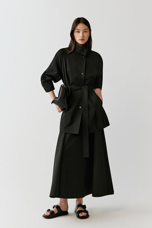 Full-length studio photograph of a woman wearing a black short trench coat in wool with a single-breasted construction. The coat features a straight cut, wide collar and relaxed shoulders and is worn over a long black skirt with flat sandals.