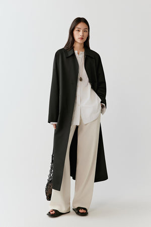 Full-length studio photograph of a woman wearing a long black trench coat in wool with a straight elongated silhouette and single-breasted construction. The coat falls below the calf and is styled over a white top and wide-leg beige trousers.