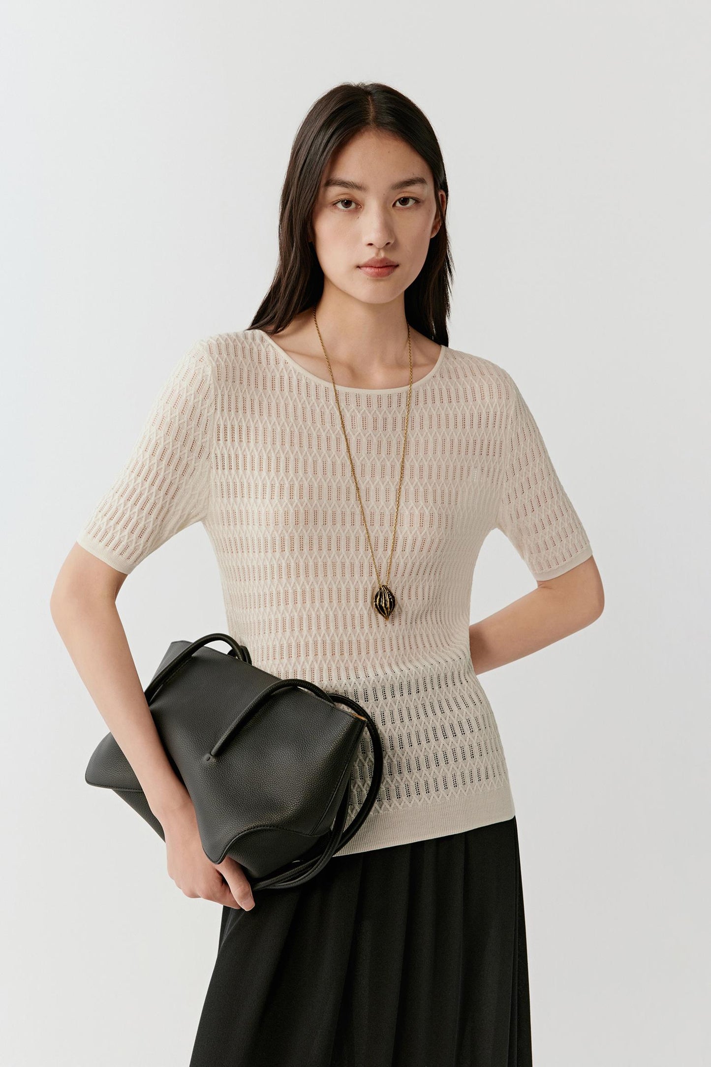 Woman wearing a cream silk and linen blend short-sleeved knit top with a straight, slightly relaxed cut and round neckline, styled with a long black skirt; front view showing the open-knit structure, breathable texture and clean shoulder line.