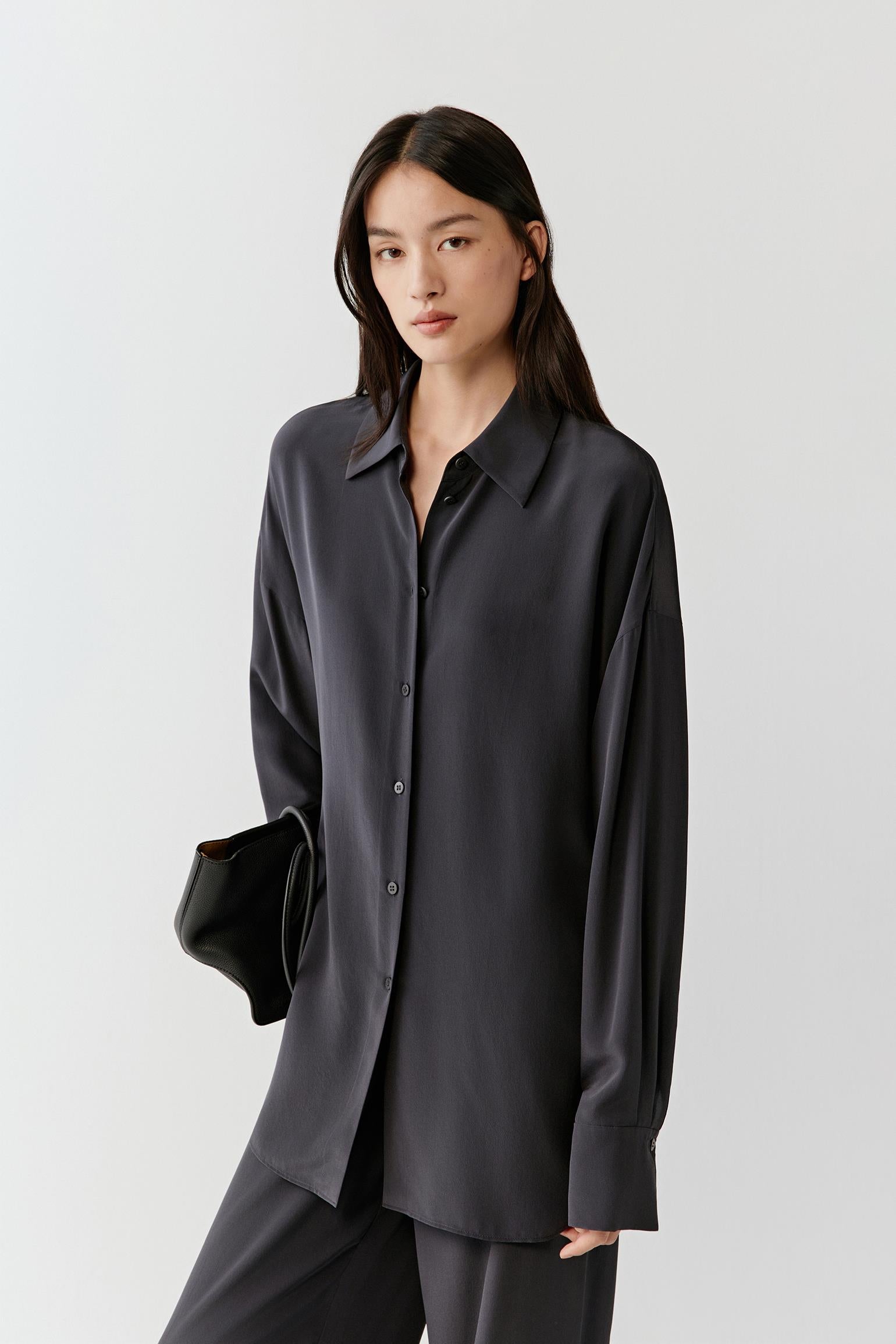 Woman wearing a black silk shirt with a relaxed, fluid cut and classic collar, front view showing the soft drape, matte finish and clean lines, styled with matching trousers.