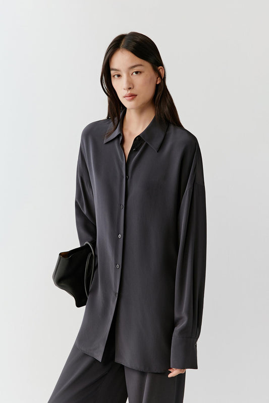 Woman wearing a black silk shirt with a relaxed, fluid cut and classic collar, front view showing the soft drape, matte finish and clean lines, styled with matching trousers.