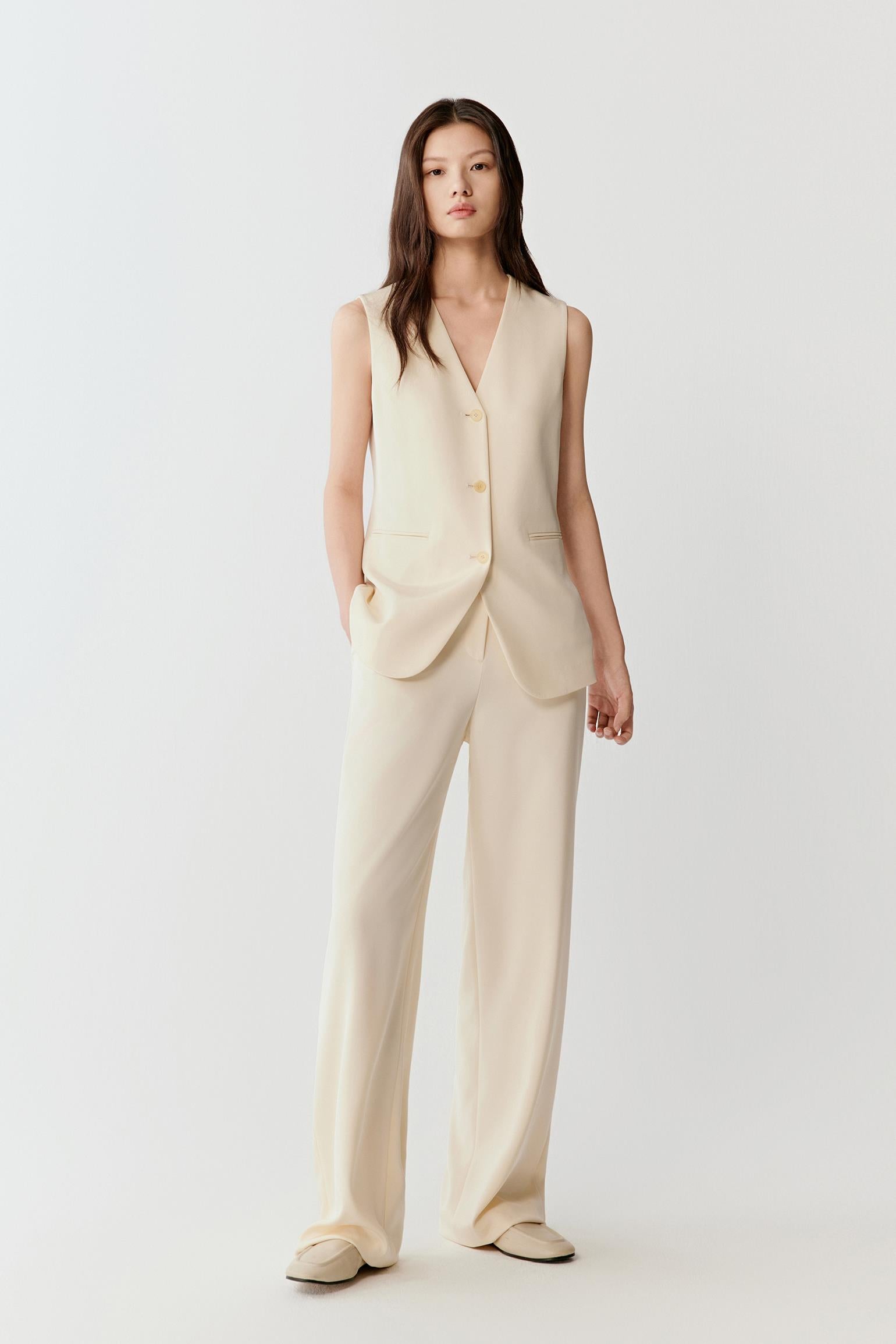 Woman wearing an ivory silk stretch heavy crepe vest jacket with a tailored, sleeveless cut and hip length, styled with matching wide-leg trousers; full-length front view showing the deep V-neckline, front button closure and fluid yet structured drape.