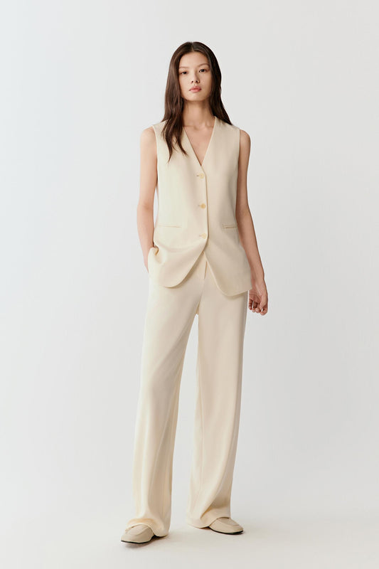 Woman wearing an ivory silk stretch heavy crepe vest jacket with a tailored, sleeveless cut and hip length, styled with matching wide-leg trousers; full-length front view showing the deep V-neckline, front button closure and fluid yet structured drape.