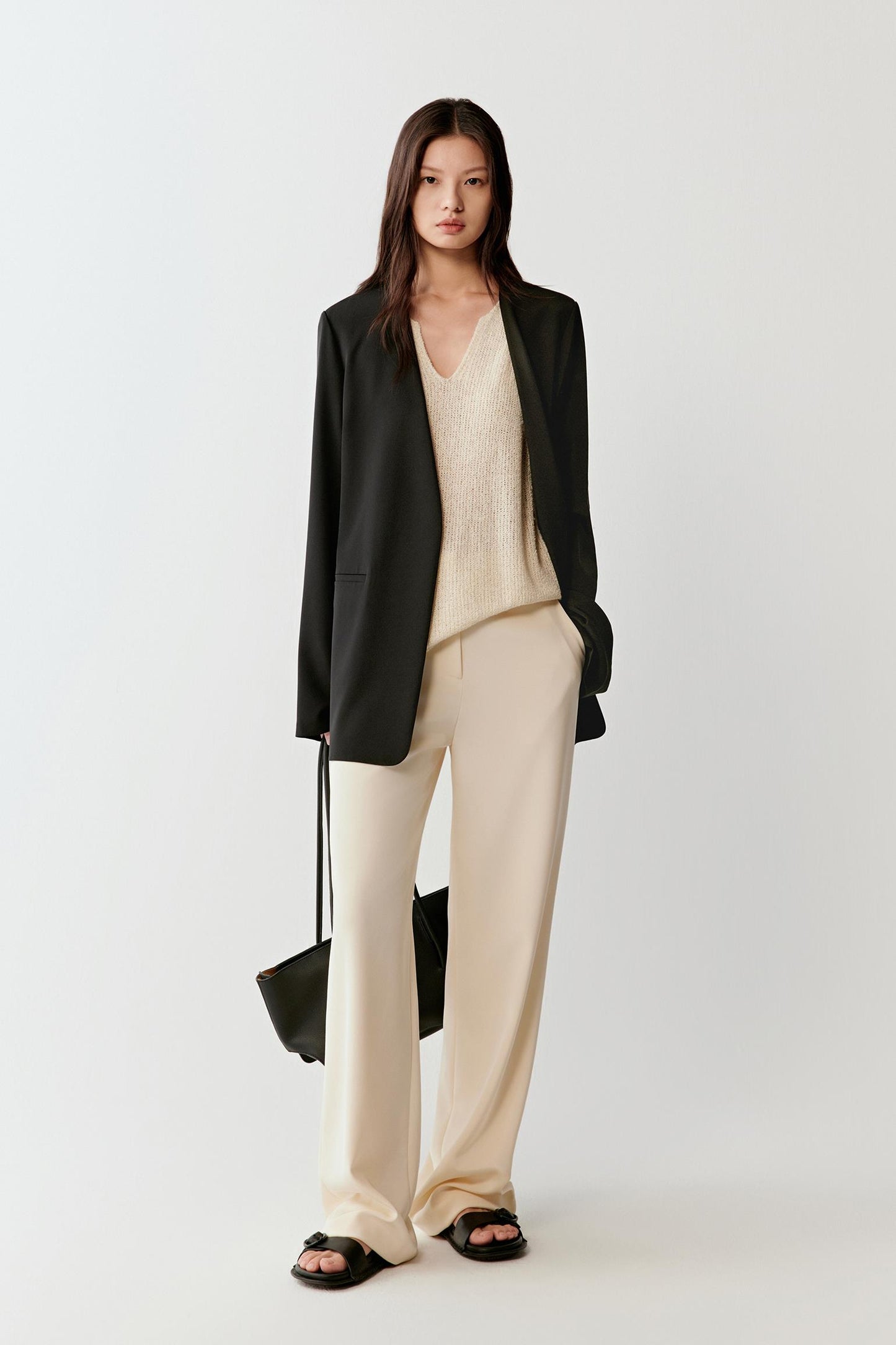 Full-length view of the black blazer paired with light beige trousers, emphasizing the long straight silhouette and contrast between dark outer layer and light inner outfit.