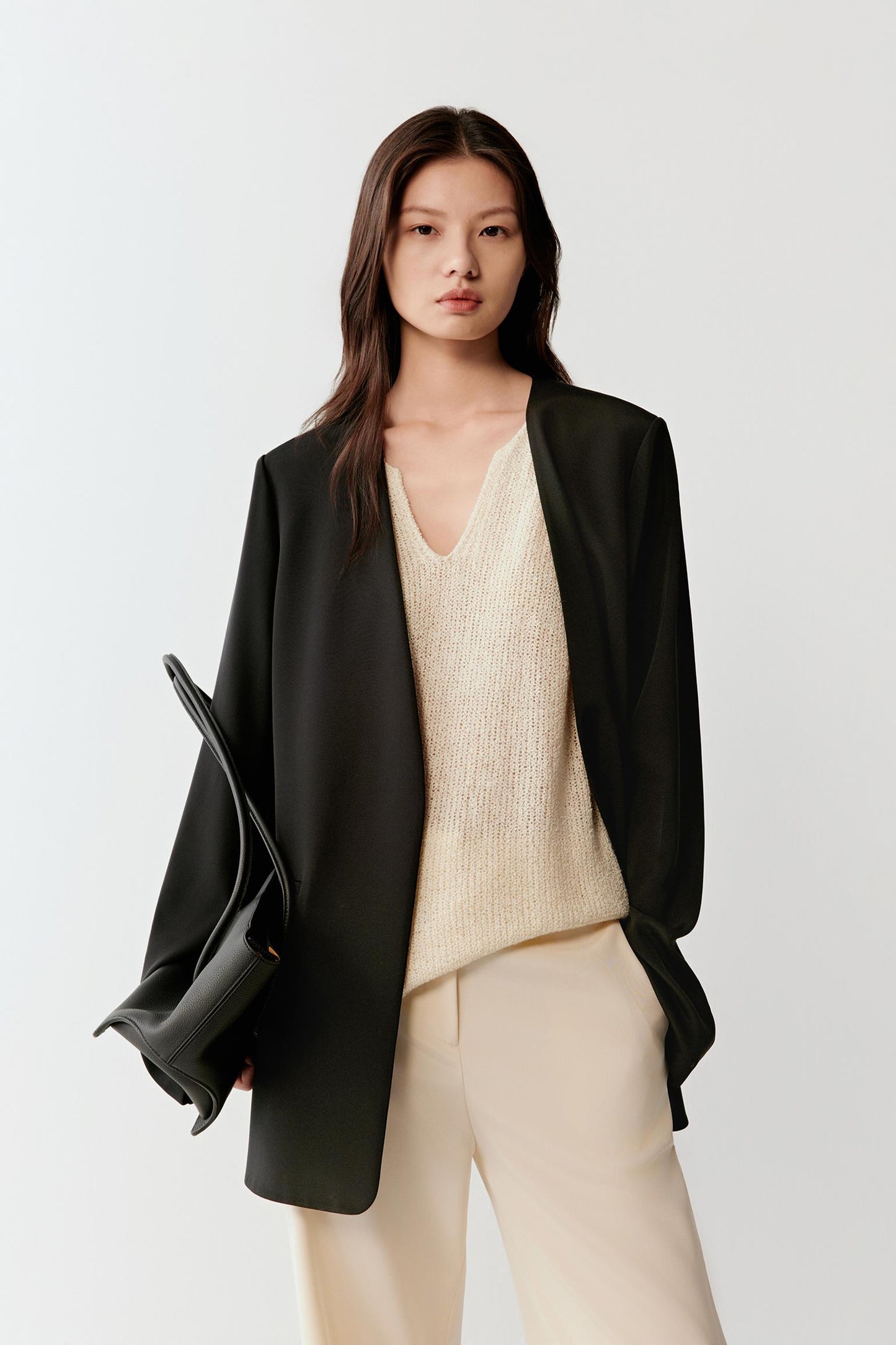 Woman wearing a black silk stretch crepe blazer with a straight, relaxed cut, styled over a light beige top, front view showing the clean lines, soft shoulders and fluid drape.