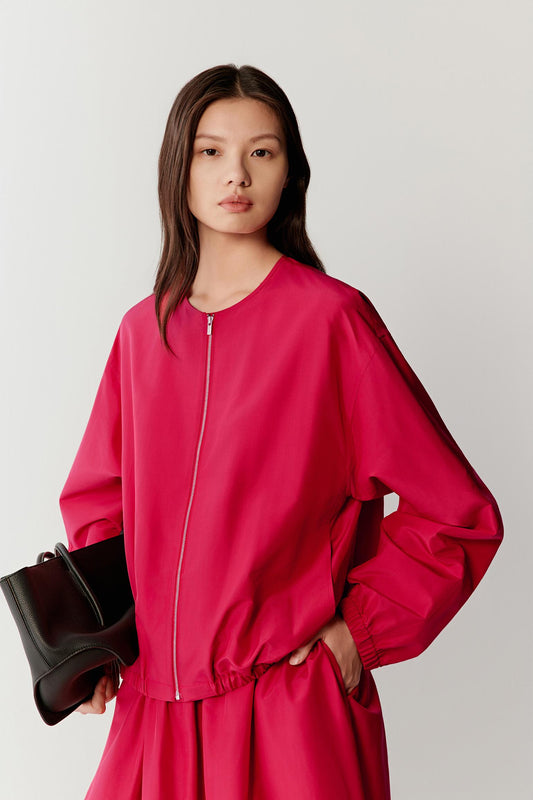 Three-quarter front view of a woman wearing a bright red silk taffeta jacket with a softly voluminous silhouette, highlighting the smooth, compact surface, structured drape and slight crispness of the fabric, with gathered volume through the sleeves and body