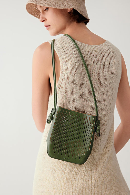 Woman wearing a khaki green perforated leather crossbody bag with a compact rectangular shape and long adjustable strap, worn across the body, back three-quarter view highlighting the openwork texture and lightweight structure.