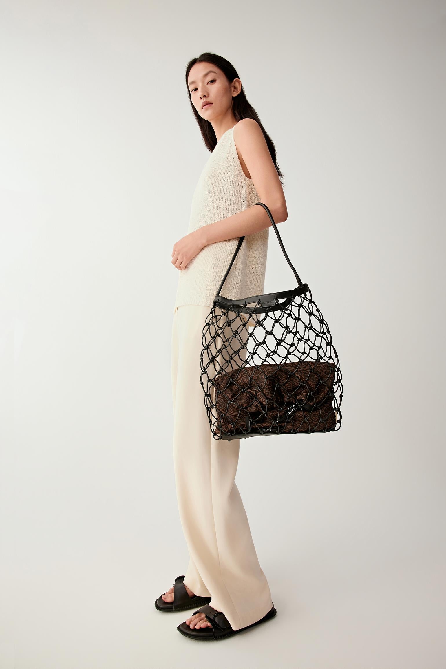 Woman wearing a black nappa leather net bag with a structured open mesh design and leather trim, carried over the shoulder, full-length view highlighting the lightweight construction and contrast with beige outfit.