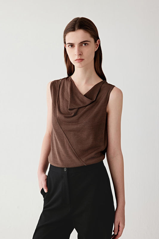 Front three-quarter view of a woman wearing a dark brown tussah silk draped top, featuring a sleeveless cut and a softly draped neckline, highlighting the fluid fall of the fabric across the chest, the natural matte texture of tussah silk and the relaxed yet structured silhouette paired with black trousers