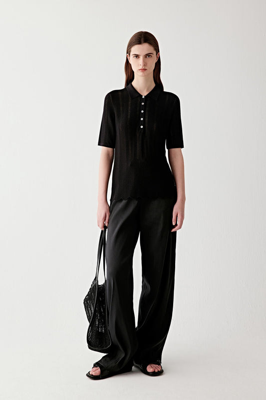 Full-length front view of a woman standing facing forward wearing a black silk and cotton knit polo, paired with wide-leg black trousers, highlighting the straight silhouette, short sleeves and the fluid yet structured drape of the openwork knit fabric