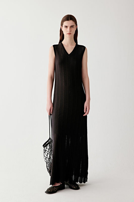 Full-length front view of a woman standing facing forward wearing a black silk and cotton knit maxi dress, featuring a sleeveless cut, deep V neckline and a long flared hem, showing a fluid silhouette, openwork knit structure and a soft, slightly sheer texture that extends to the ankles