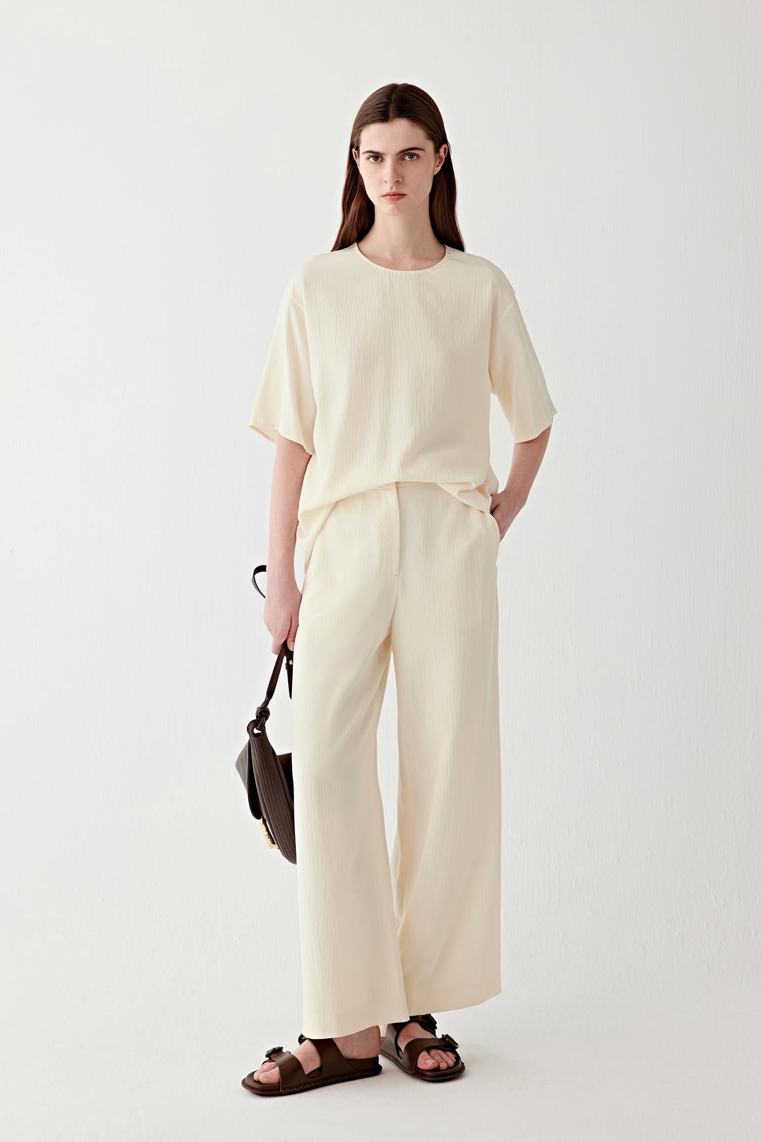 Full-length front view of a woman standing facing forward wearing an ivory silk blouse with short sleeves, paired with matching wide-leg trousers, highlighting the fluid drape, relaxed silhouette and softly crinkled texture of the lightweight silk fabric