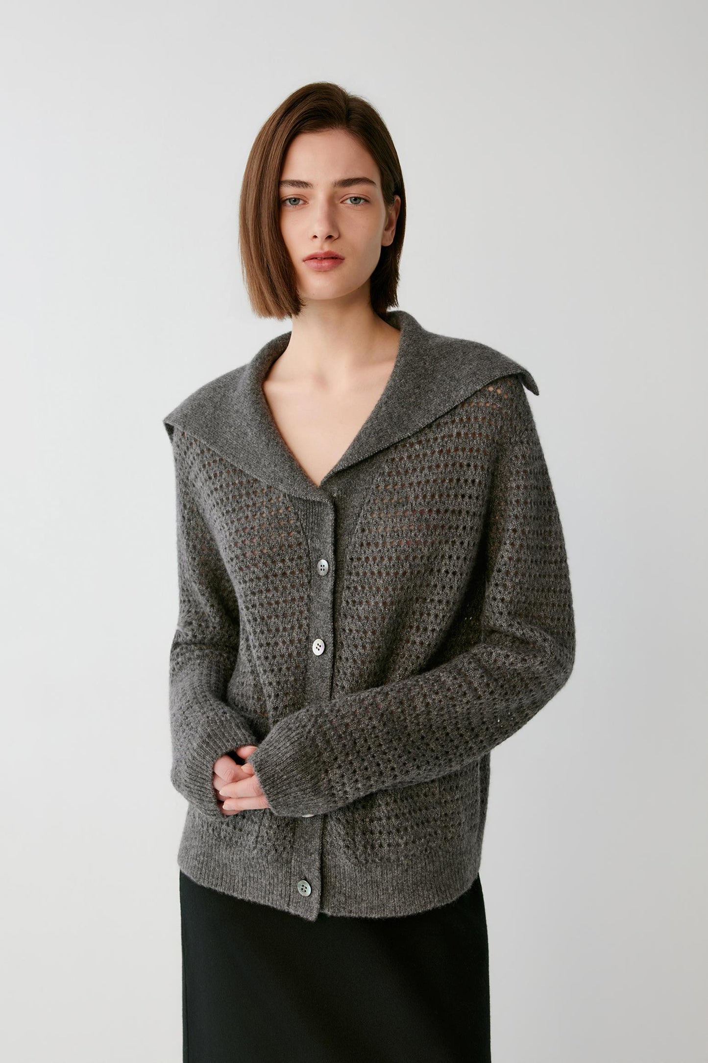 A woman wearing a textured gray knit cardigan with a wide collar and button front, paired with a black skirt, photographed against a plain light gray studio background.