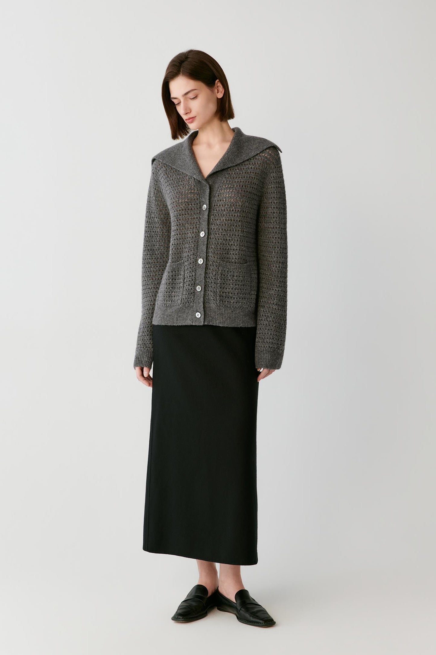 A model stands against a light gray studio background wearing a textured gray cropped cardigan with a wide collar and front buttons, paired with a long black skirt and black loafers.
