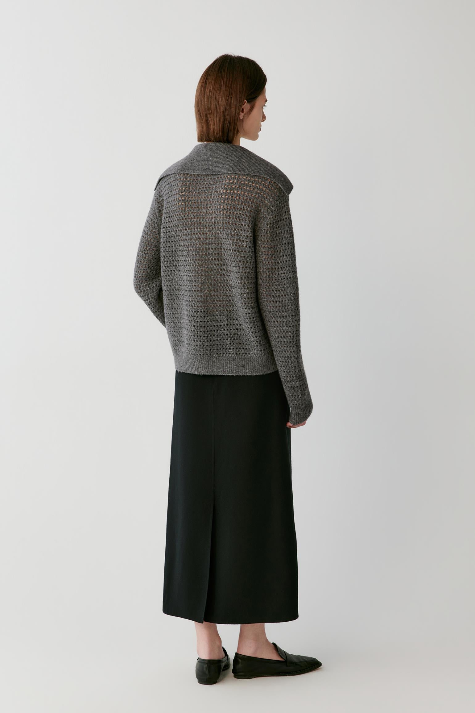 A model is shown from the back wearing a gray open-knit cropped cardigan and a long black skirt, standing against a light gray studio background.
