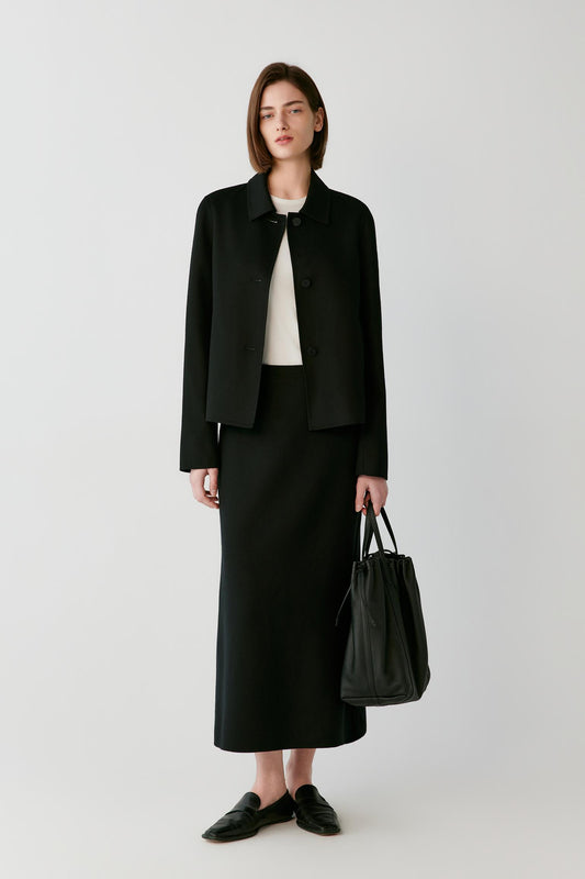 A model stands against a light gray studio background wearing a black cropped jacket, black midi skirt, black loafers, and holding a black leather tote bag.