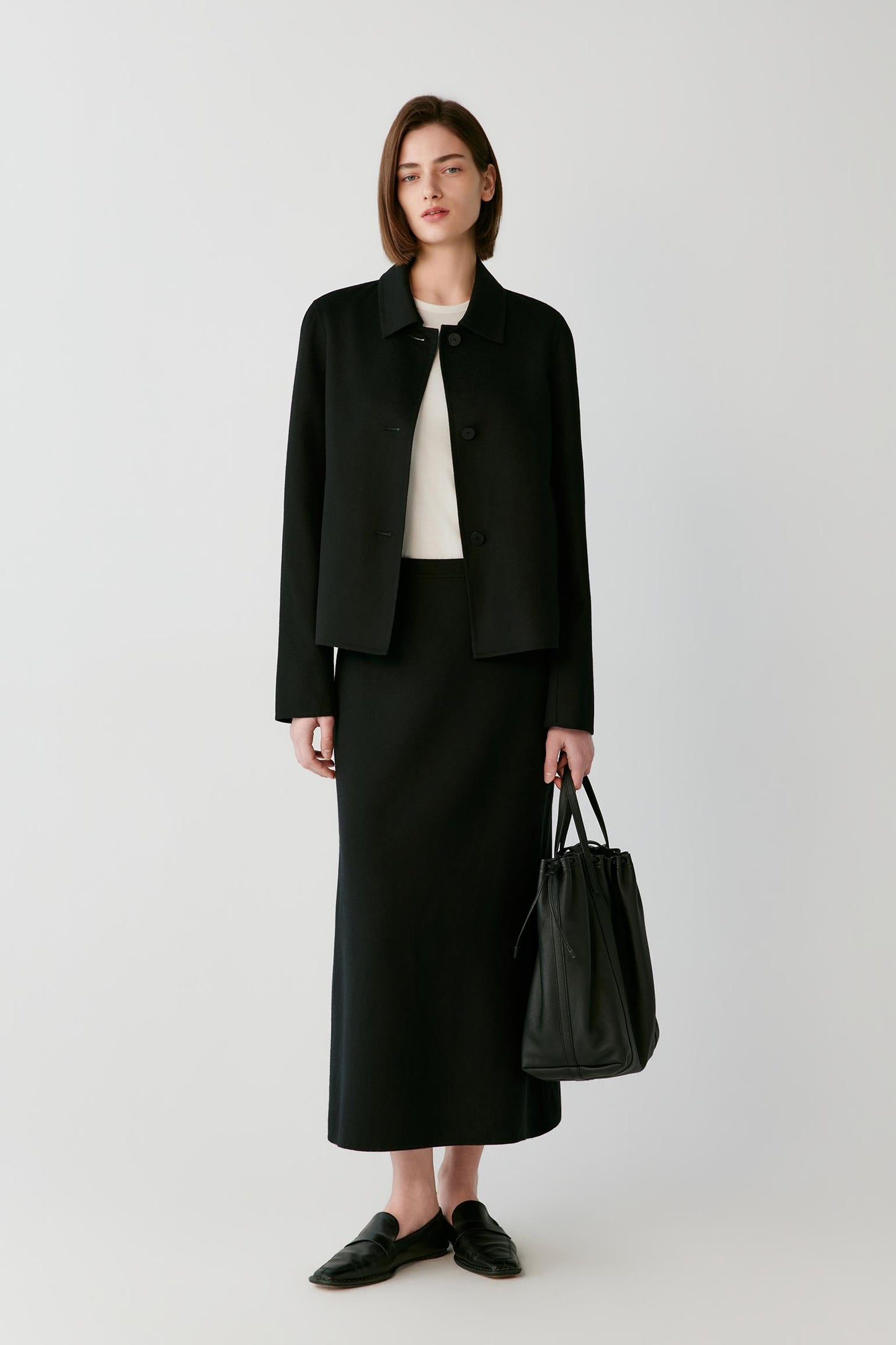A model stands against a light gray studio background wearing a black cropped jacket, black midi skirt, black loafers, and holding a black leather tote bag.