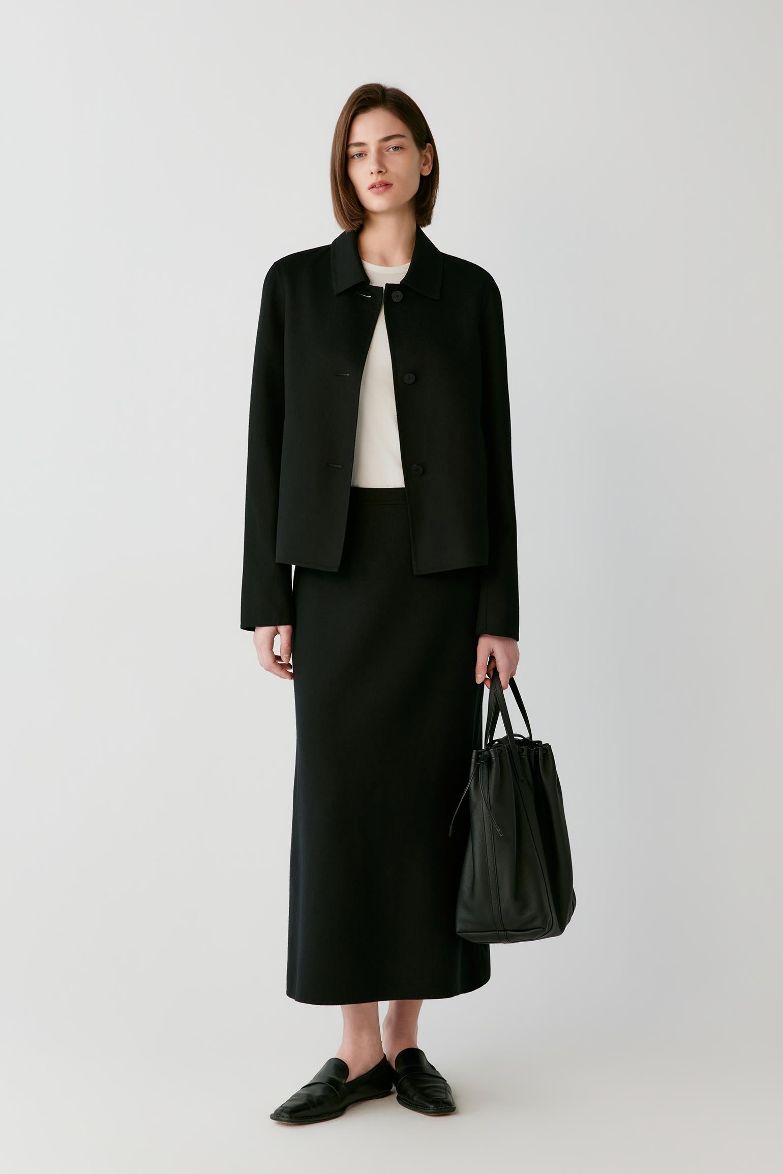 A model stands against a light gray studio background wearing a black cropped jacket, black midi skirt, black loafers, and holding a black leather tote bag.