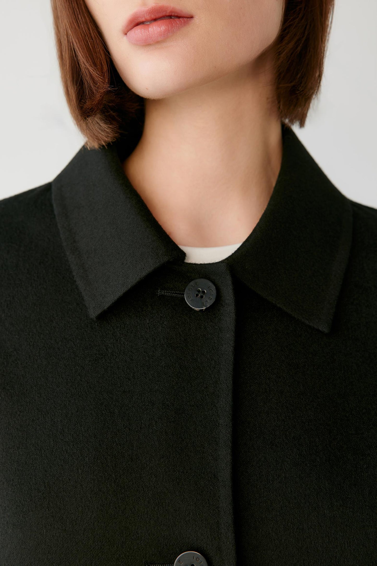 Close-up of a model wearing a black collared coat with a single dark button, over a white inner layer, against a light gray background.