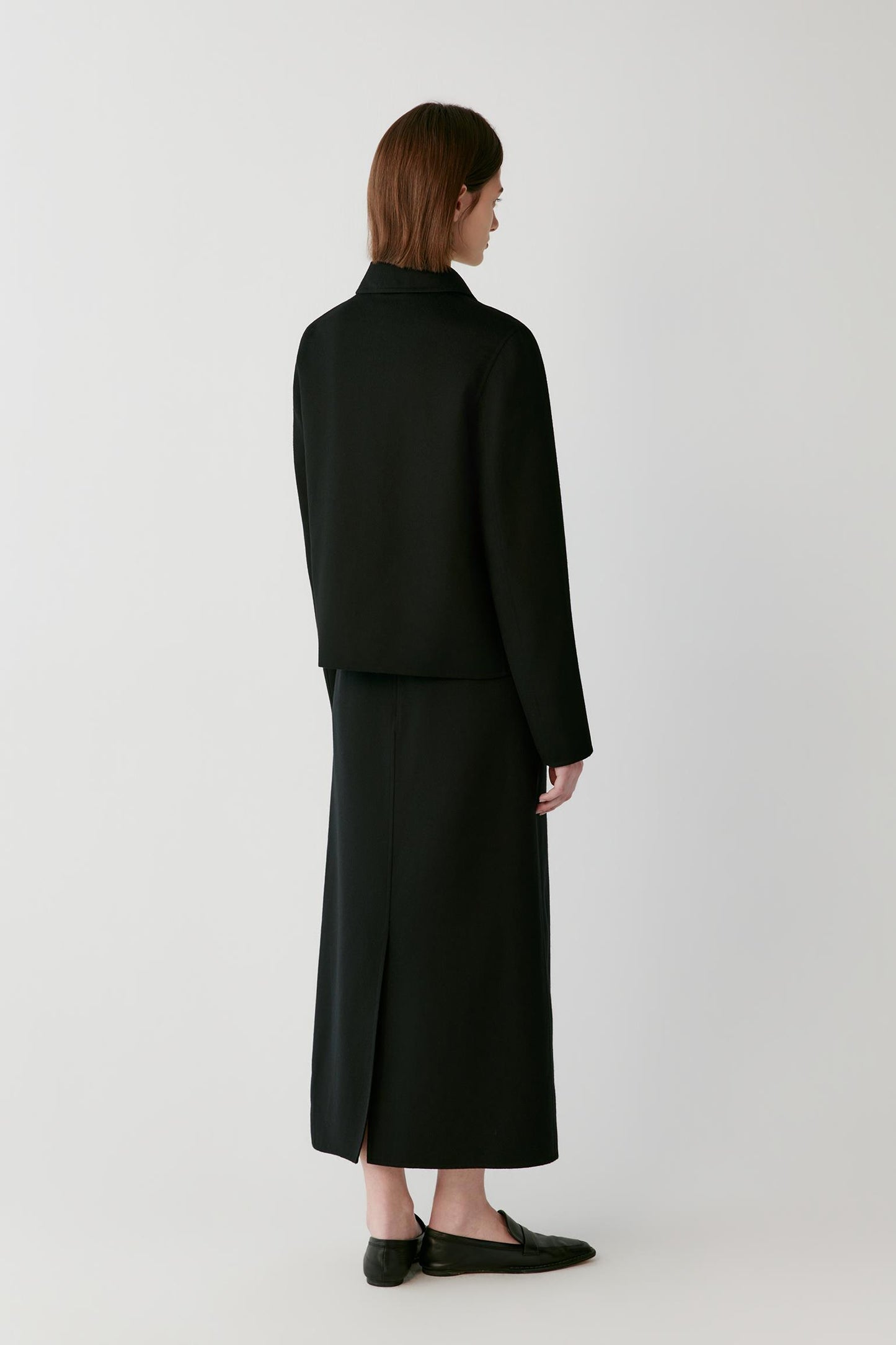 A model stands against a light gray studio background wearing a black cropped jacket, black midi skirt, black loafers, and holding a black leather tote bag.