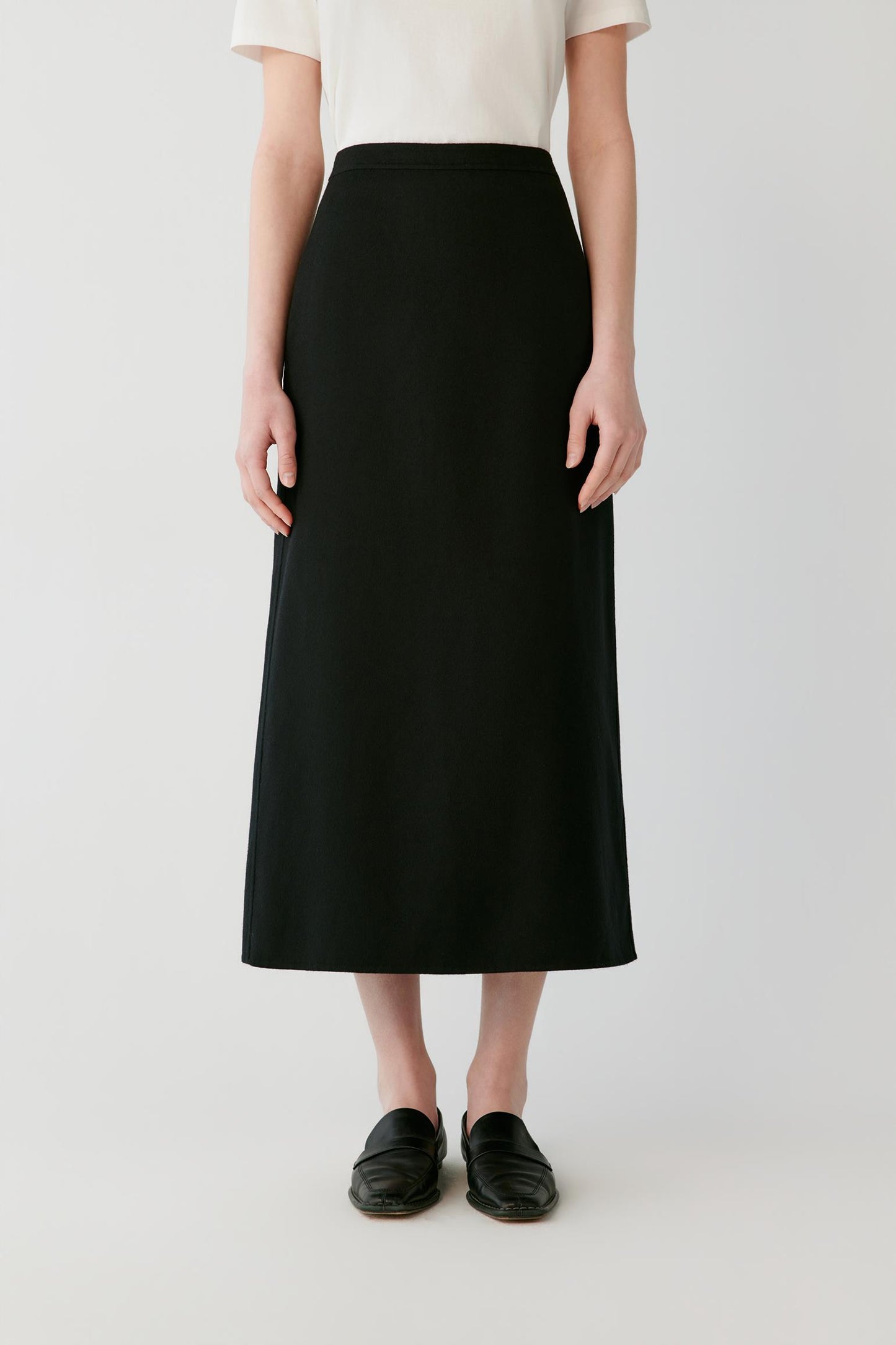 A model wearing a high-waisted, black midi skirt paired with a cream short-sleeve top and black leather loafers, photographed against a light gray studio background.