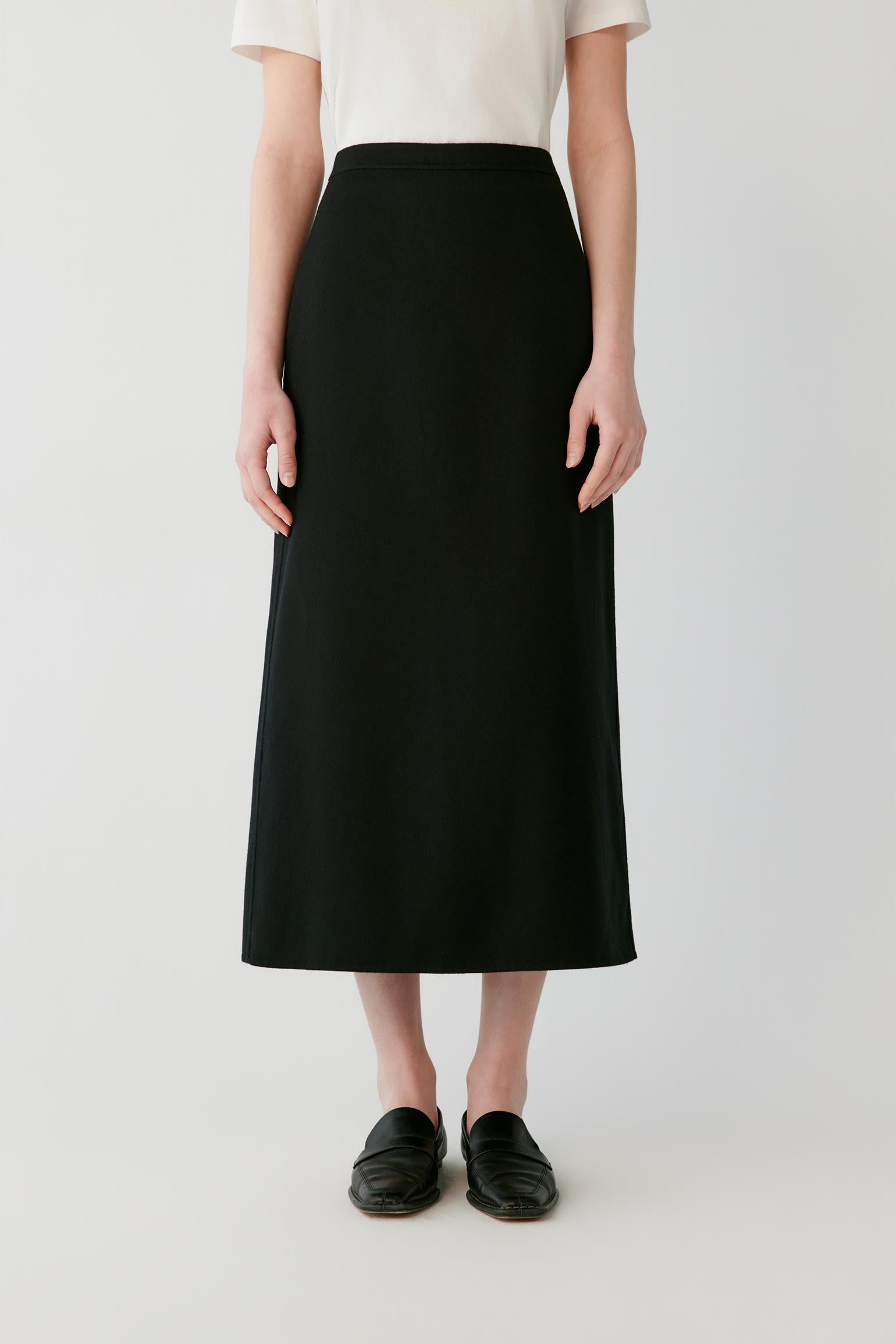 A model wearing a high-waisted, black midi skirt paired with a cream short-sleeve top and black leather loafers, photographed against a light gray studio background.