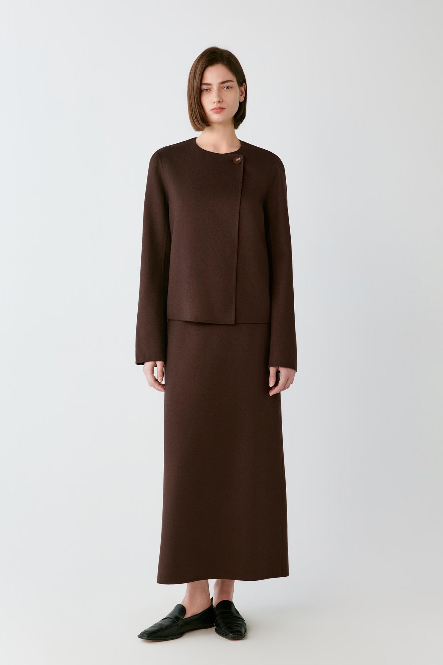 A model stands against a light gray studio background wearing a dark brown two-piece outfit consisting of a cropped long-sleeve jacket with an asymmetrical button detail and a matching midi skirt, paired with black loafers.
