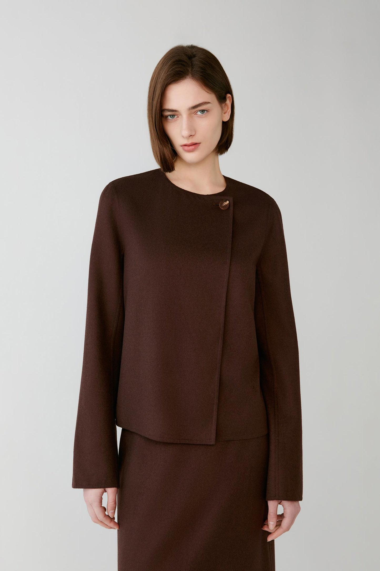 A woman modeling a dark brown wrap-style jacket with a single decorative button detail at the collar, paired with a matching brown skirt, against a light gray studio background.