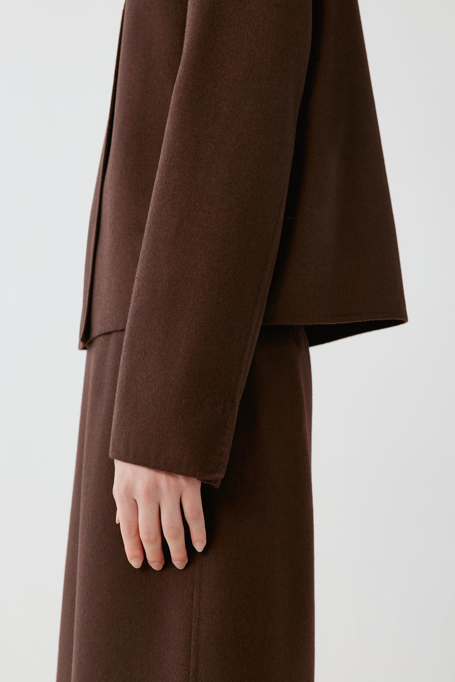 A close-up side view of a person wearing a dark brown wool coat with a matching long skirt or dress, showing the sleeve, hand, and drape of the fabric against a plain light gray background.