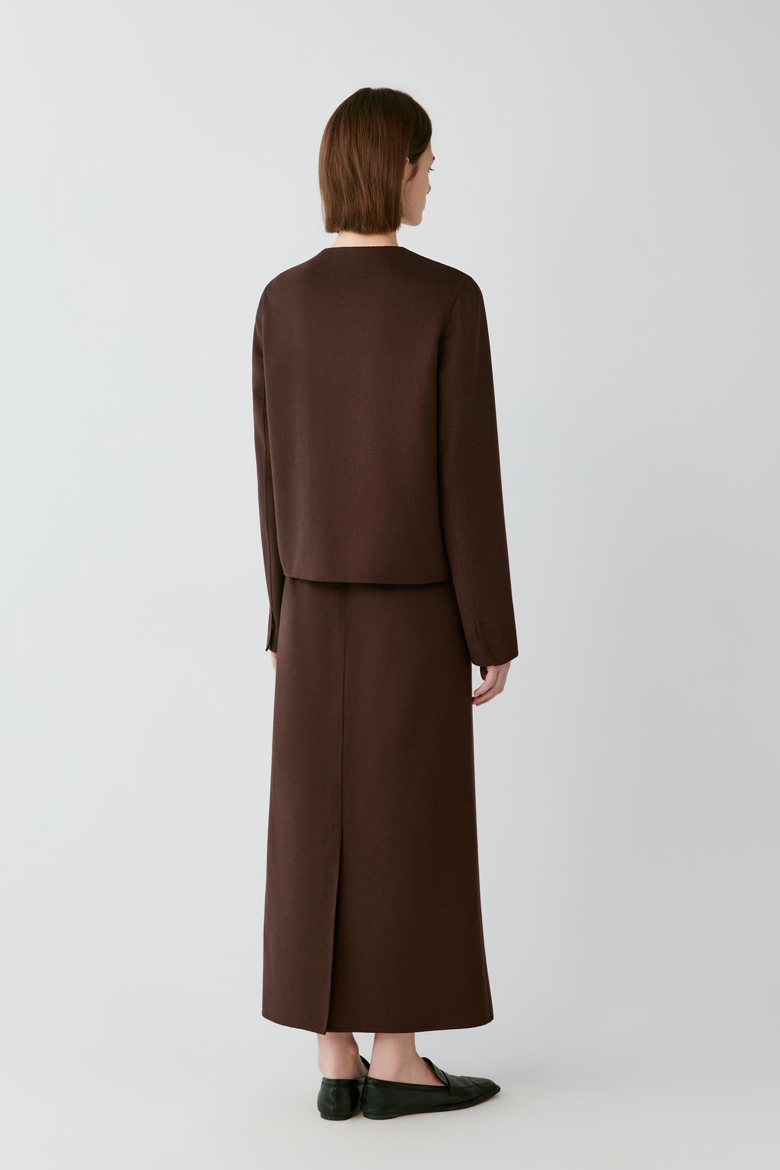 A model shown from the back wearing a dark brown two-piece outfit consisting of a cropped long-sleeve jacket and a matching midi skirt, paired with black flat shoes against a light gray studio background.