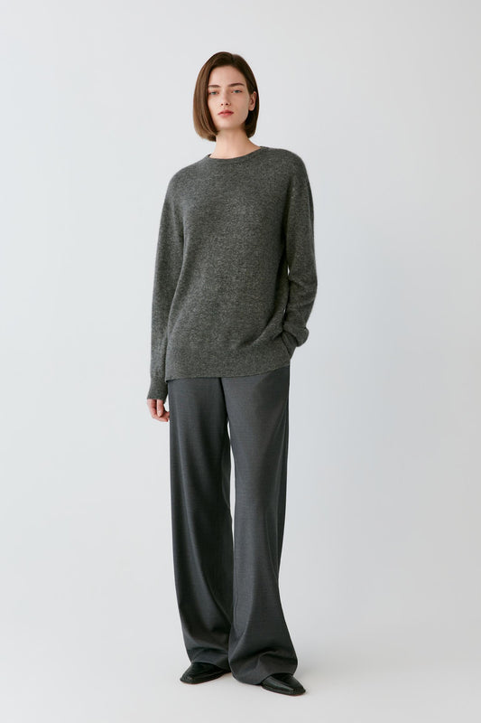 A model stands against a light gray studio background wearing a dark gray knit crewneck sweater and matching dark gray wide-leg trousers.