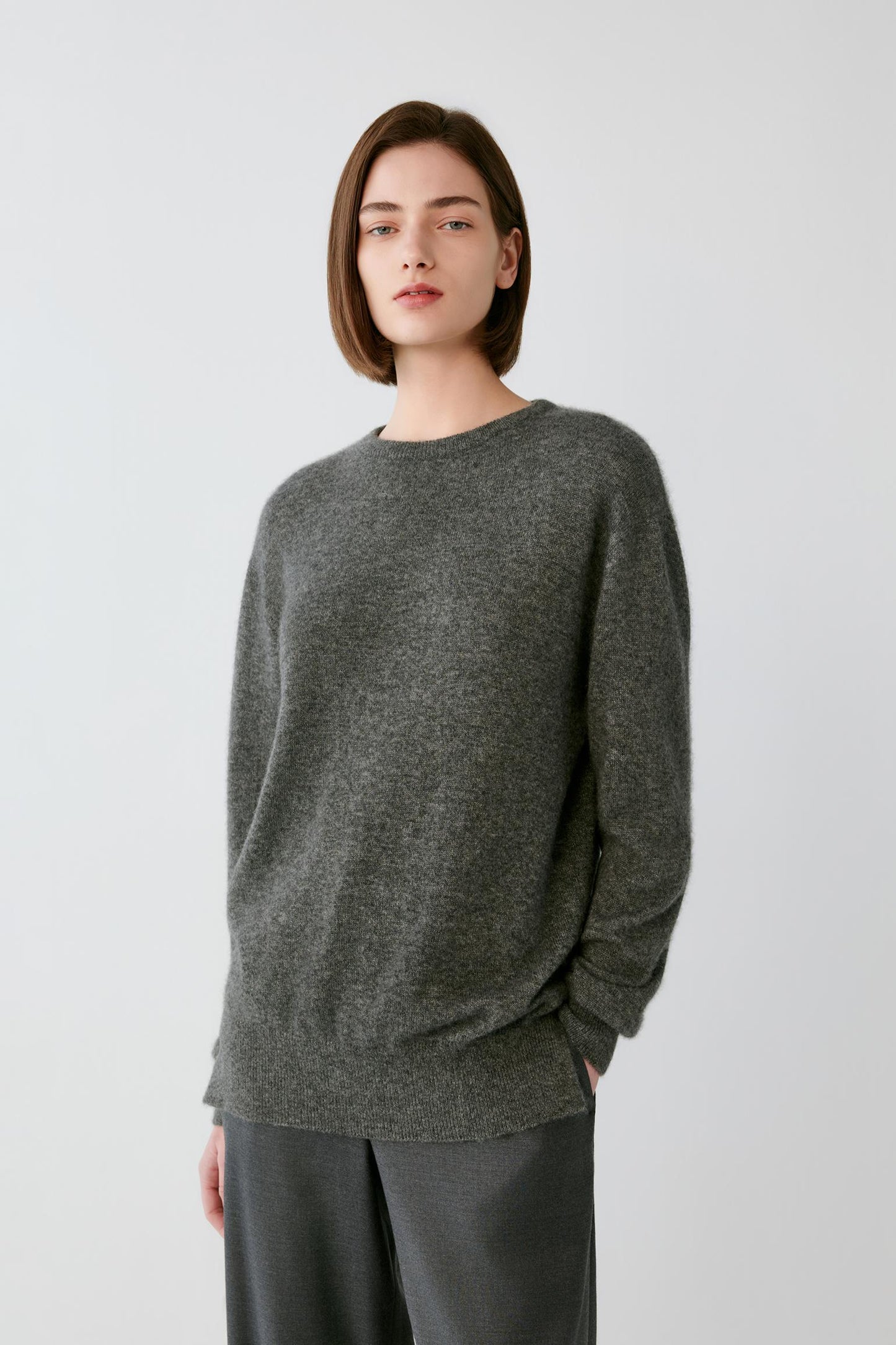 A woman with short brown hair wearing a dark gray heathered knit sweater and matching dark gray trousers, standing against a light gray studio background.