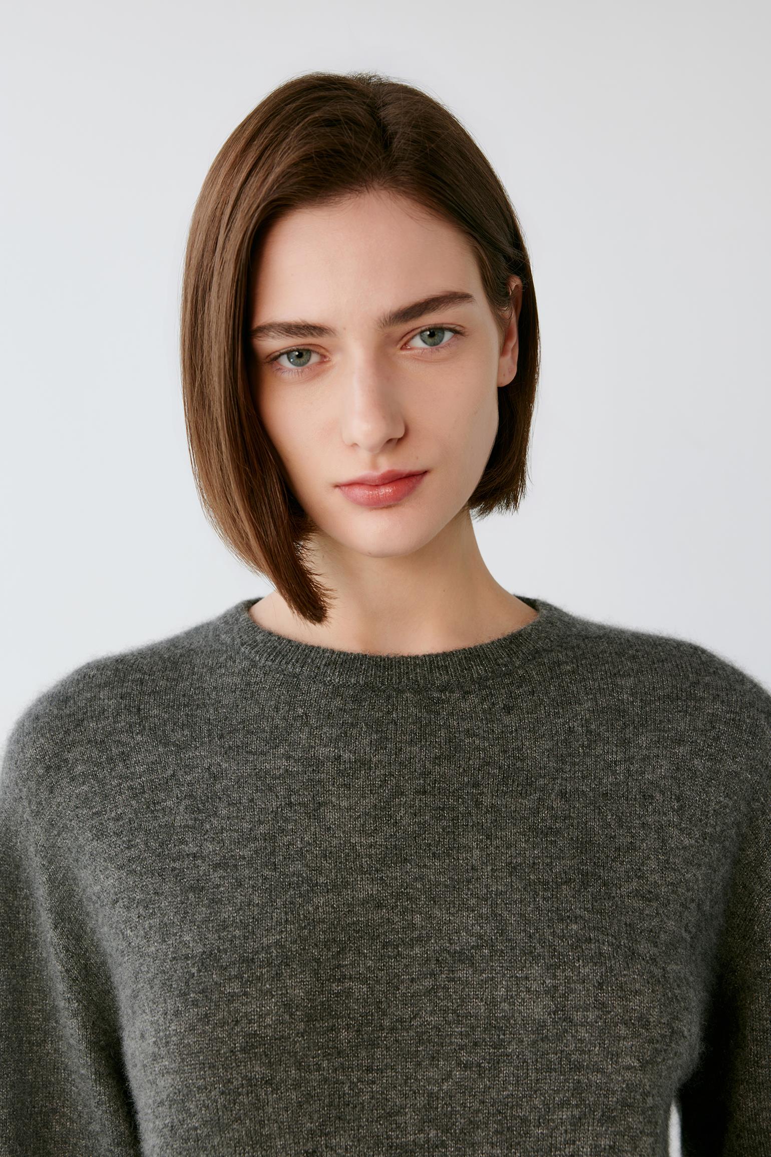 A woman with a brown bob haircut wearing a dark gray knit crewneck sweater, photographed against a light gray studio background.
