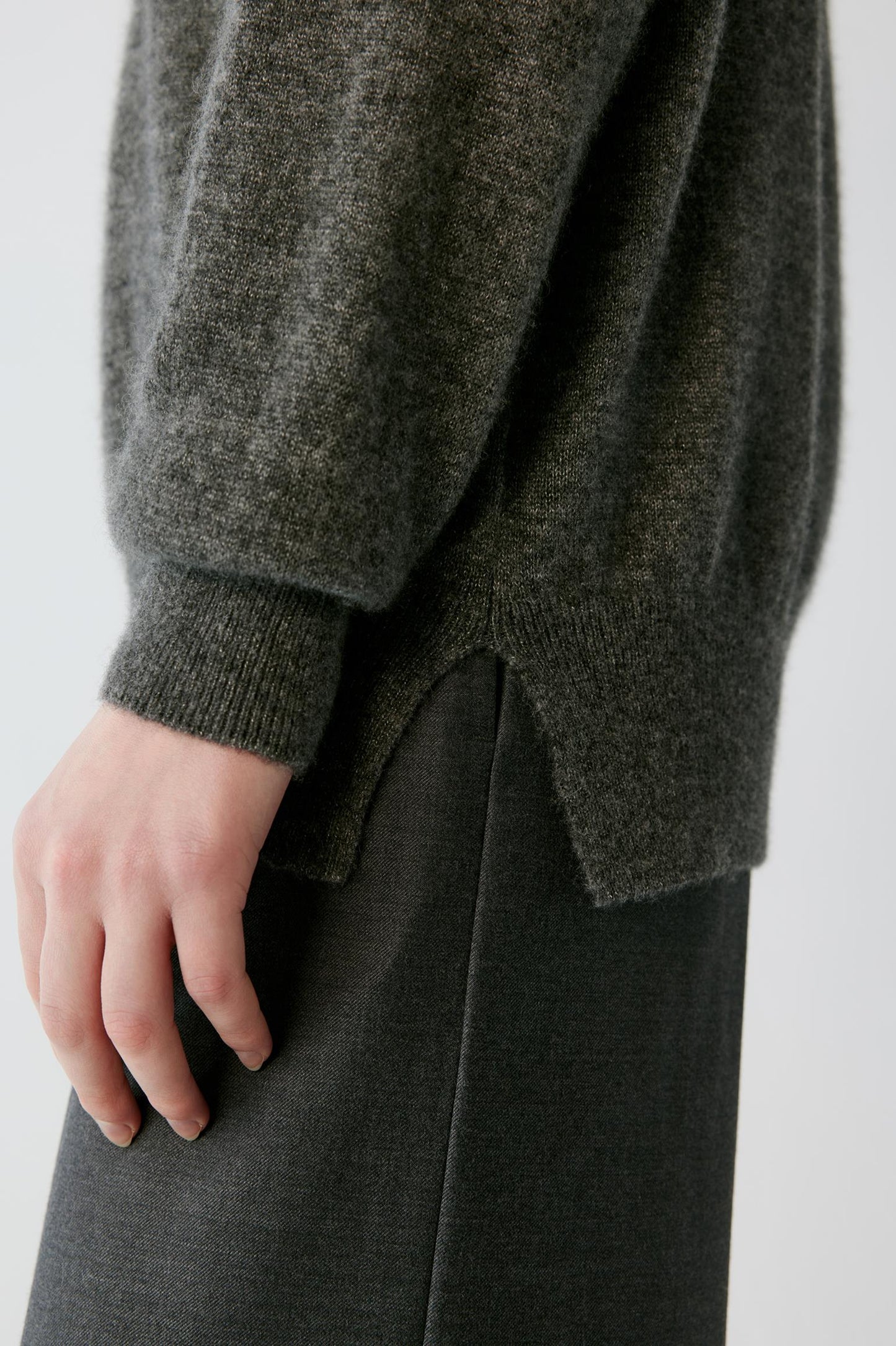 Close-up of a person wearing a dark olive green knit sweater with a ribbed cuff and hem, paired with a dark skirt, against a light gray background.