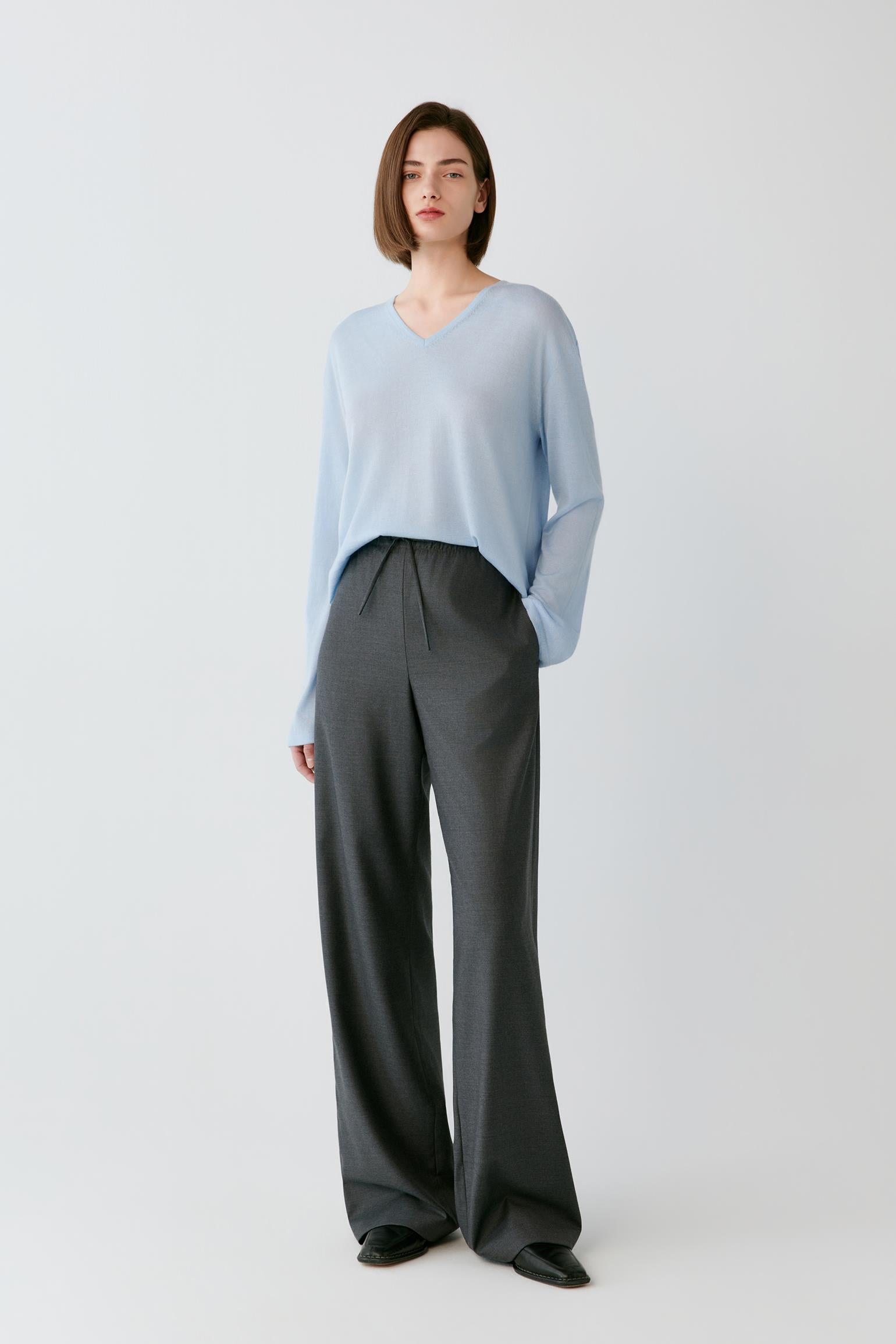 A model stands against a light gray studio background wearing a light blue long-sleeve V-neck top and dark charcoal gray wide-leg trousers.