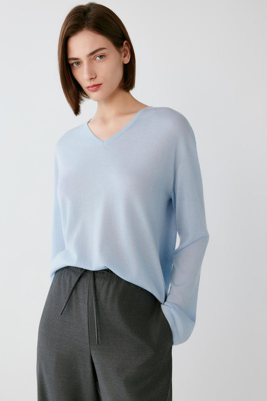 A woman wearing a light blue long-sleeve V-neck knit top and dark gray drawstring trousers, photographed against a plain light gray studio background.