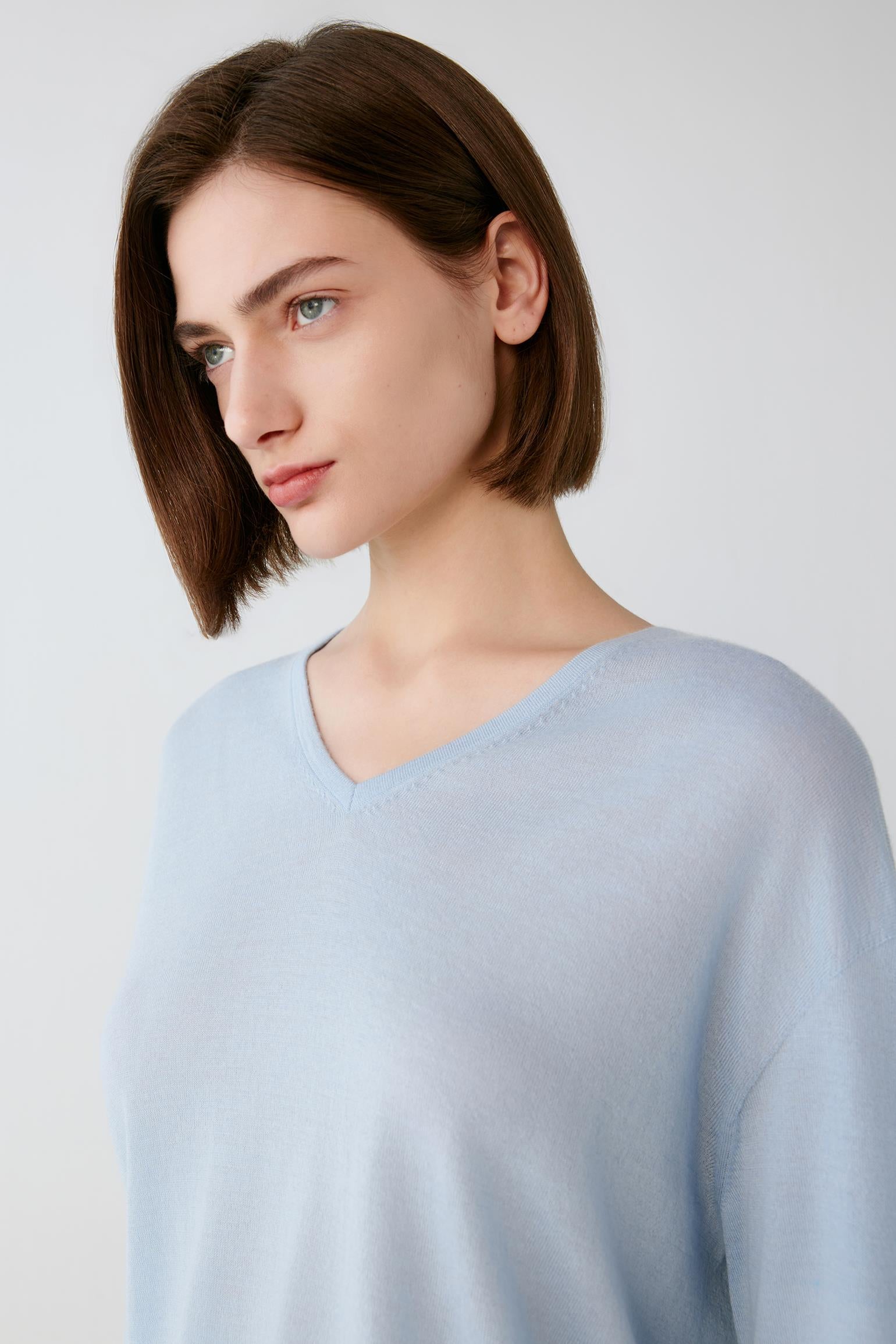 A woman with a short brown bob haircut wearing a light blue V-neck knit sweater, photographed against a plain light gray studio background.