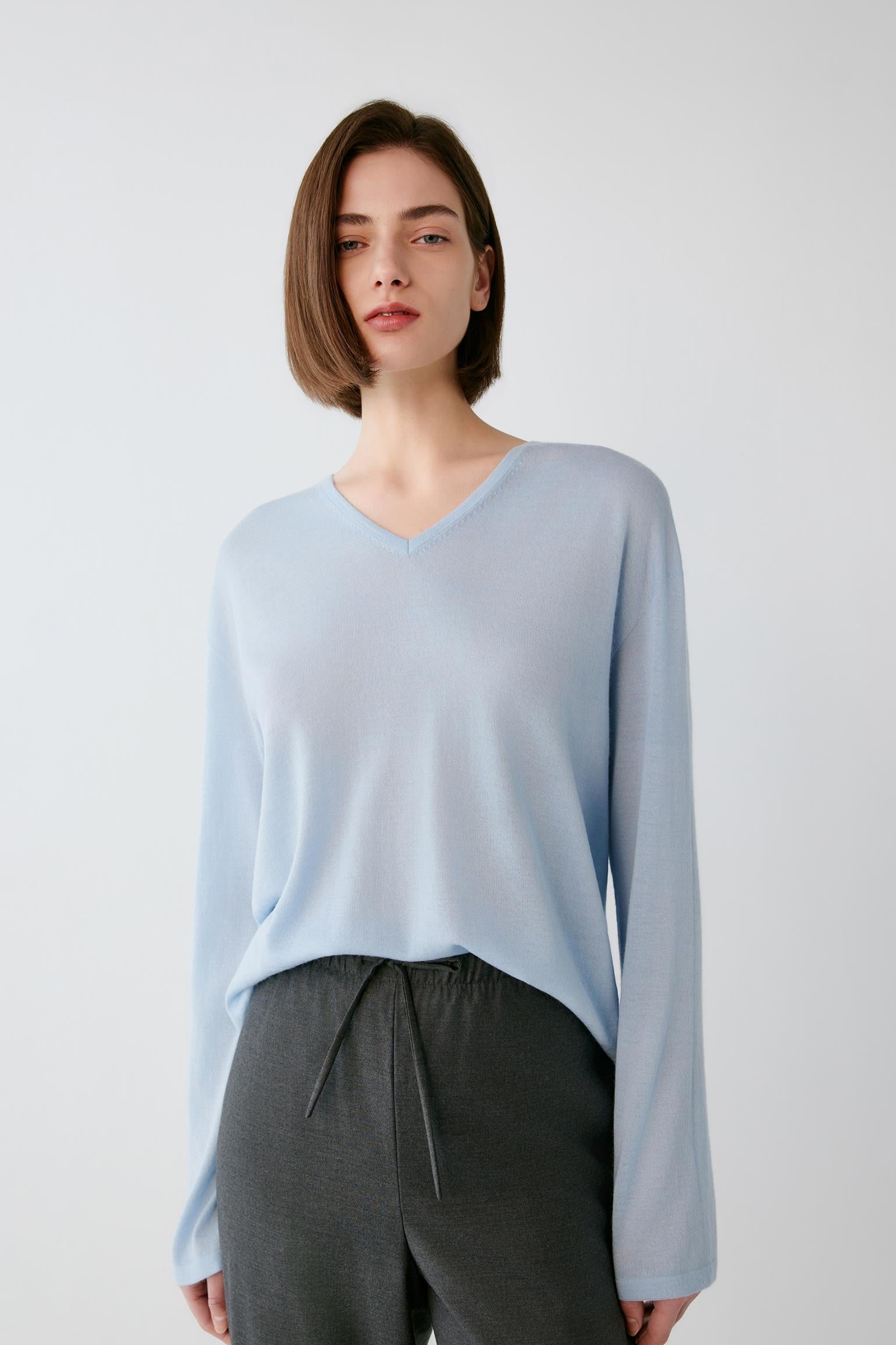 A woman wearing a light blue long-sleeve V-neck knit top and dark gray drawstring pants, standing against a plain light gray studio background.