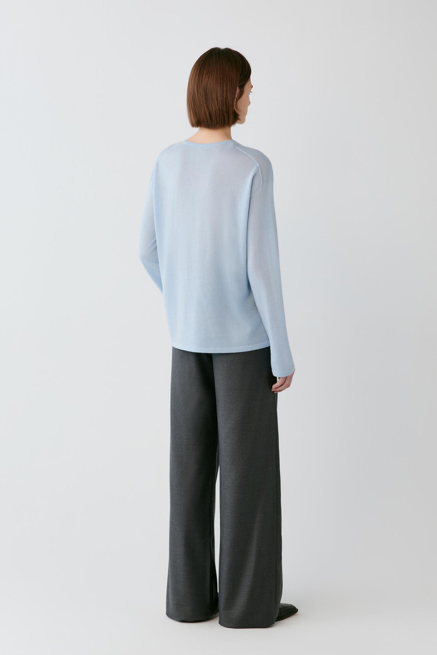 A model shown from the back wearing a light blue long-sleeve knit top and dark gray wide-leg trousers, standing against a plain light gray studio background.