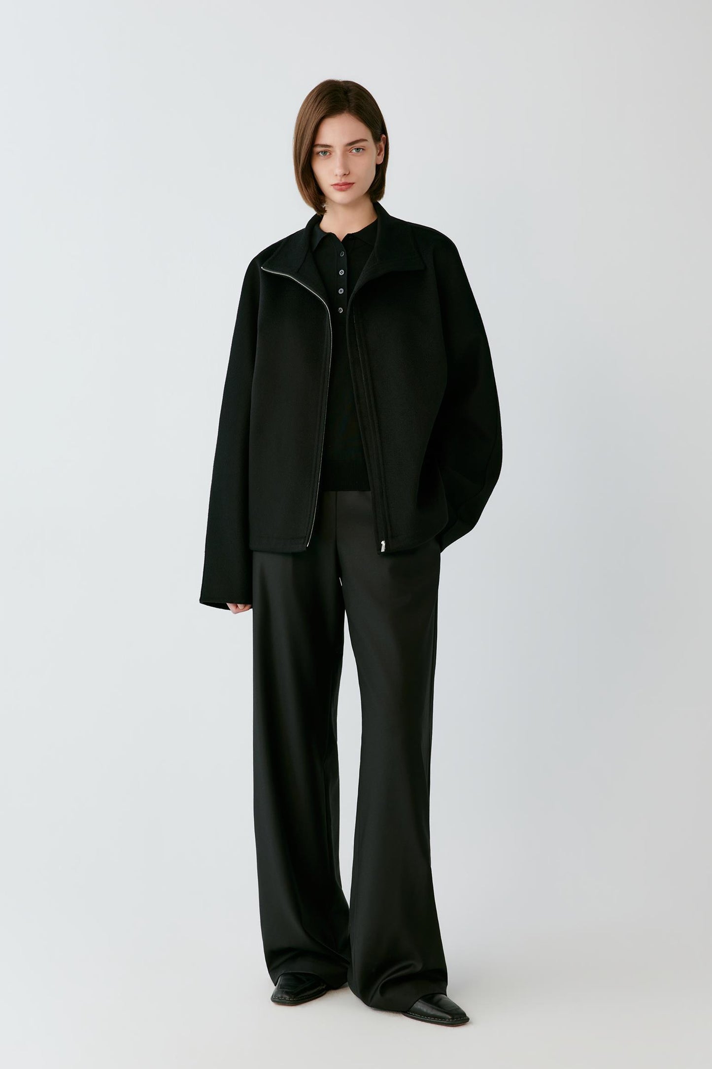 A model wearing a black zip-up jacket with a high mock neck collar, photographed against a neutral light gray background.