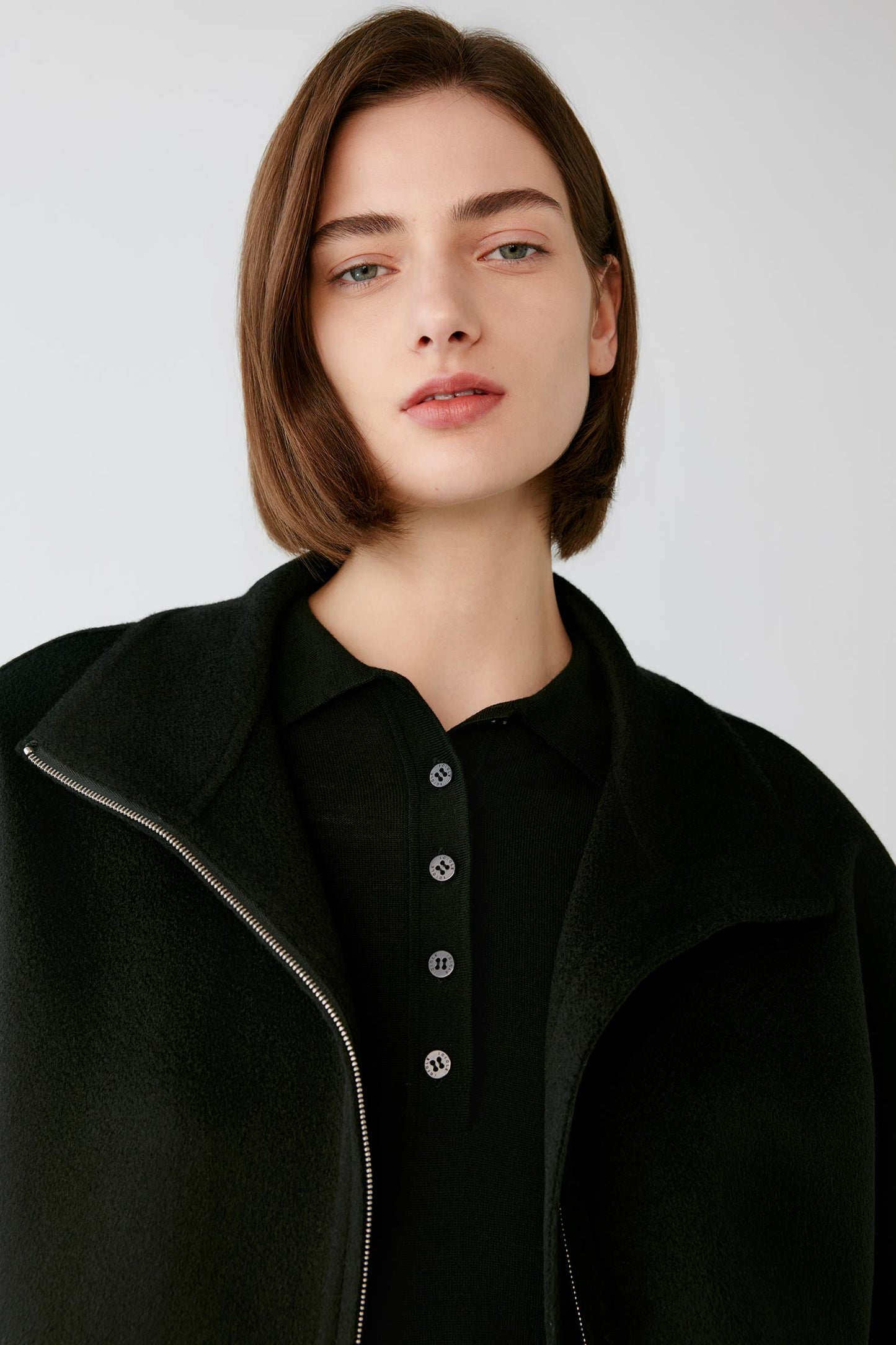 A woman with a short brown bob haircut wearing a black zip-up coat over a black buttoned collared shirt, photographed against a light gray studio background.