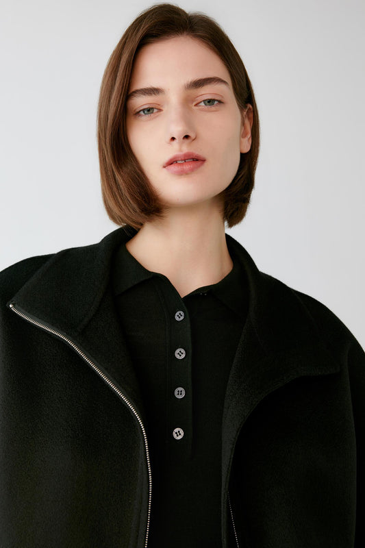 A woman with a short brown bob haircut wearing a black zip-up coat over a black buttoned collared shirt, photographed against a light gray studio background.