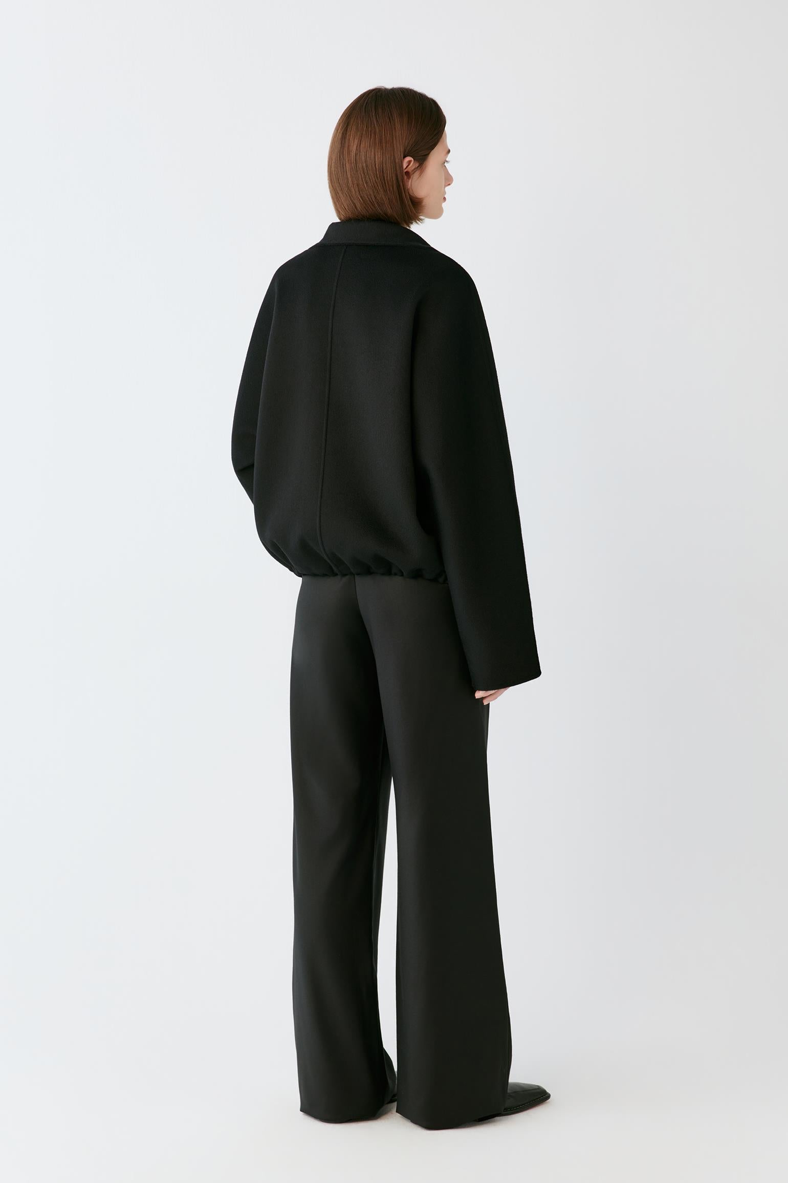 A model shown from the back wearing a black long-sleeve collared jacket and matching wide-leg trousers against a plain light gray studio background.