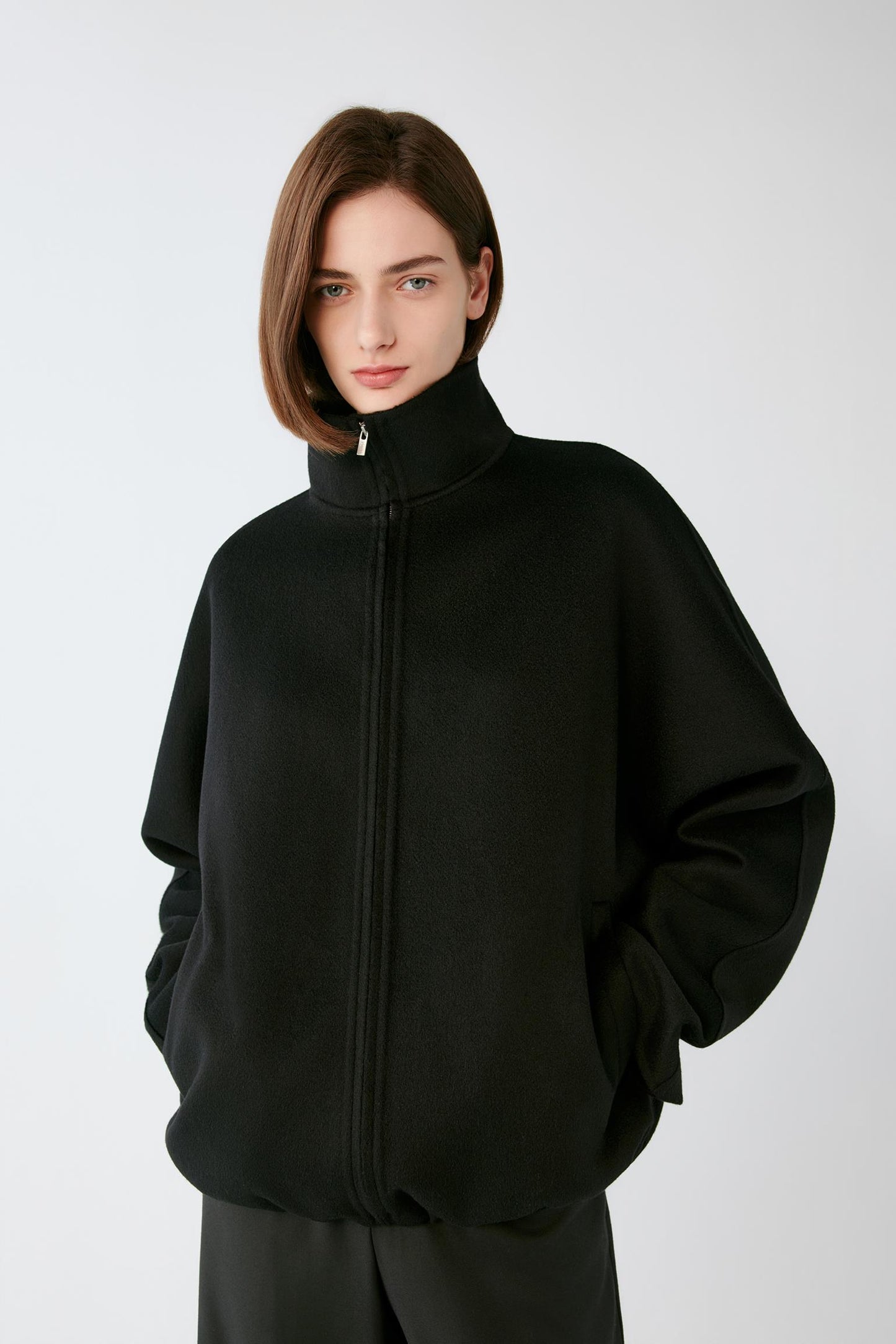 A model wearing a black zip-up jacket with a high mock neck collar, photographed against a neutral light gray background.