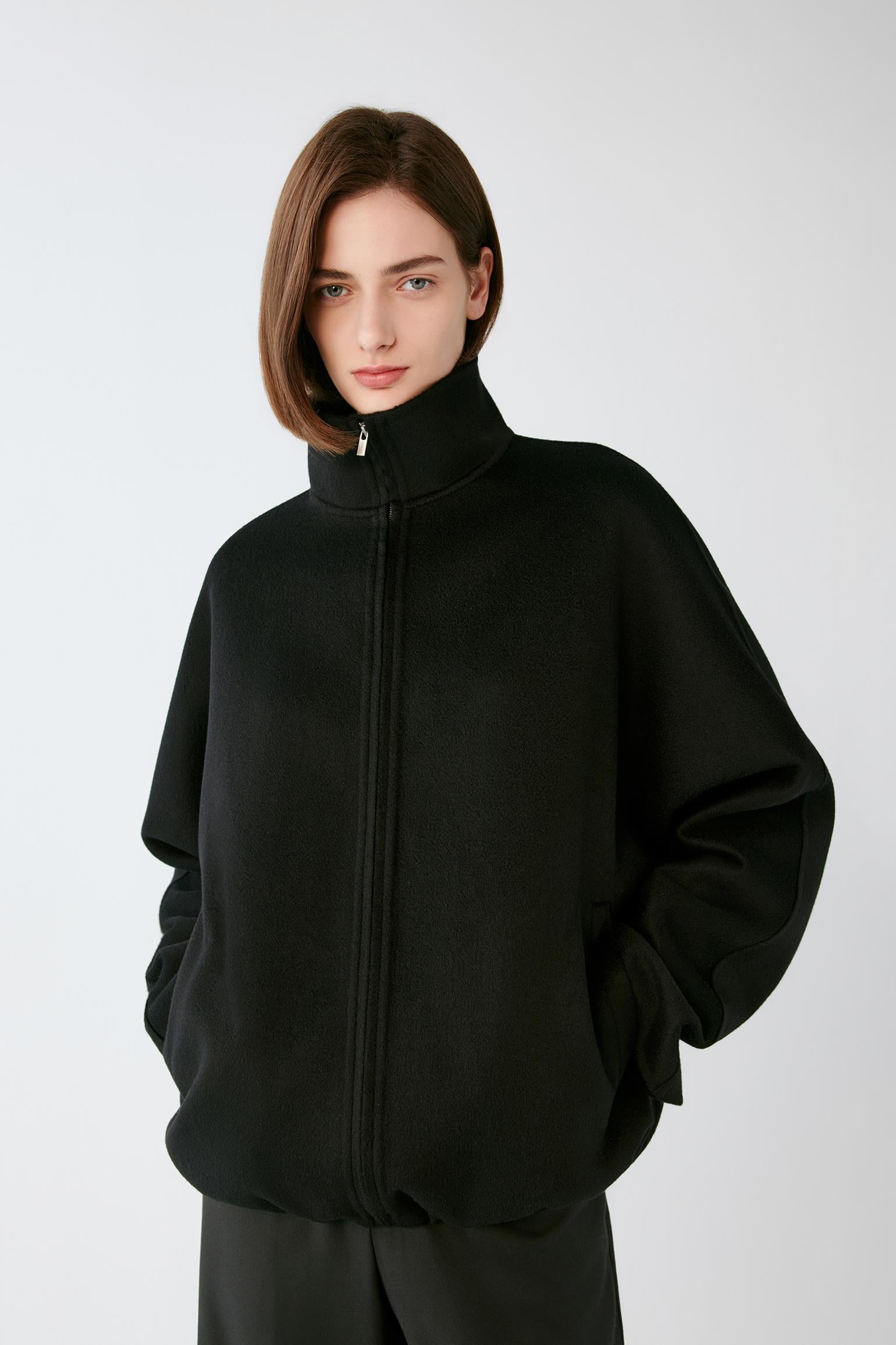 A model wearing a black zip-up jacket with a high mock neck collar, photographed against a neutral light gray background.