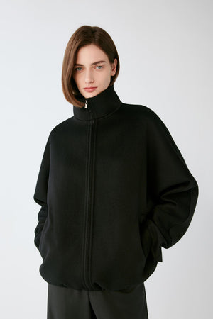 A model wearing a black zip-up jacket with a high mock neck collar, photographed against a neutral light gray background.